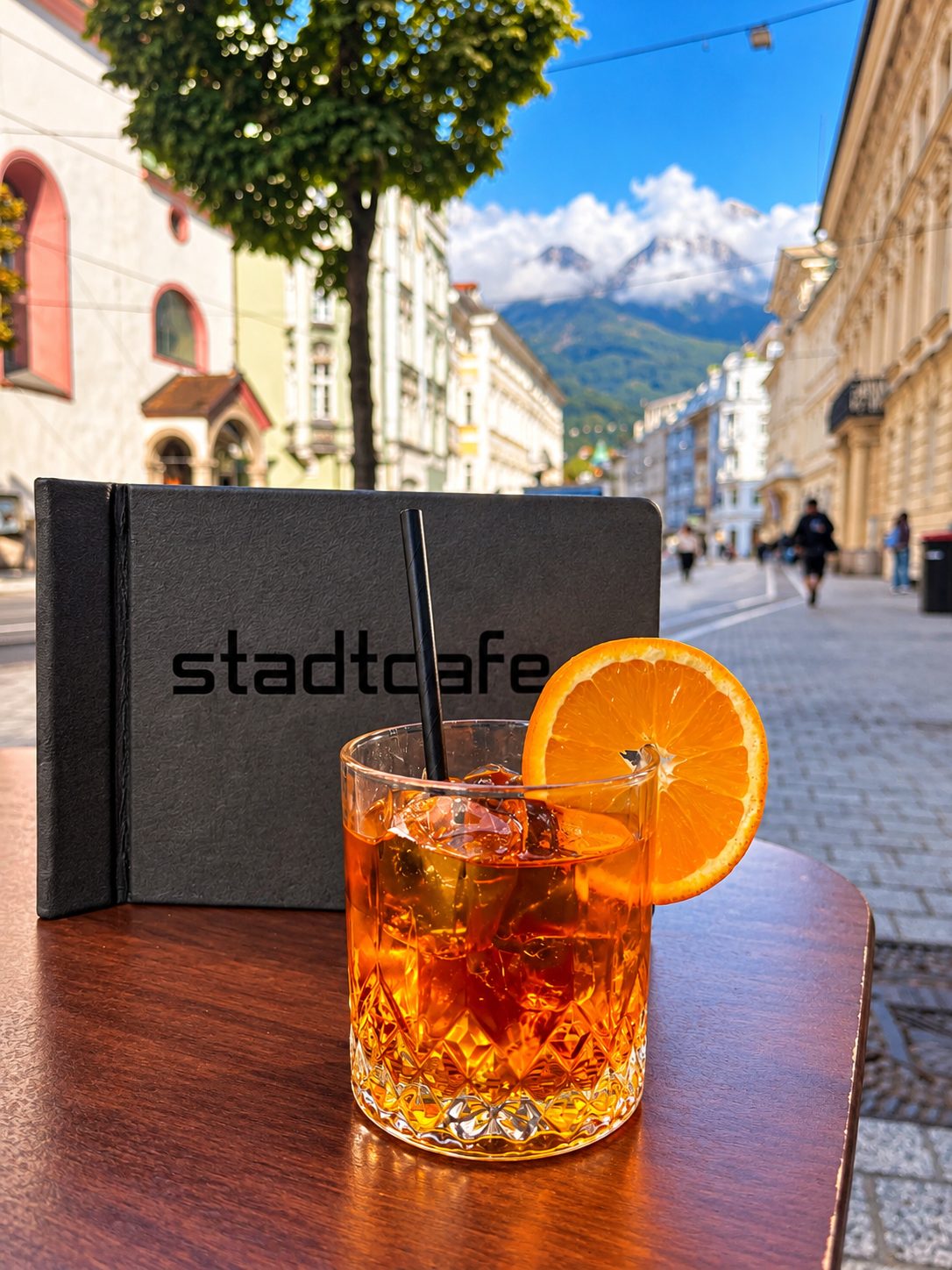 Ein Aperitivo mit Eis und Orangenscheibe auf einem Tisch vor dem Café, dahinter die Maria-Theresien-Straße und die verschneite Nordkette