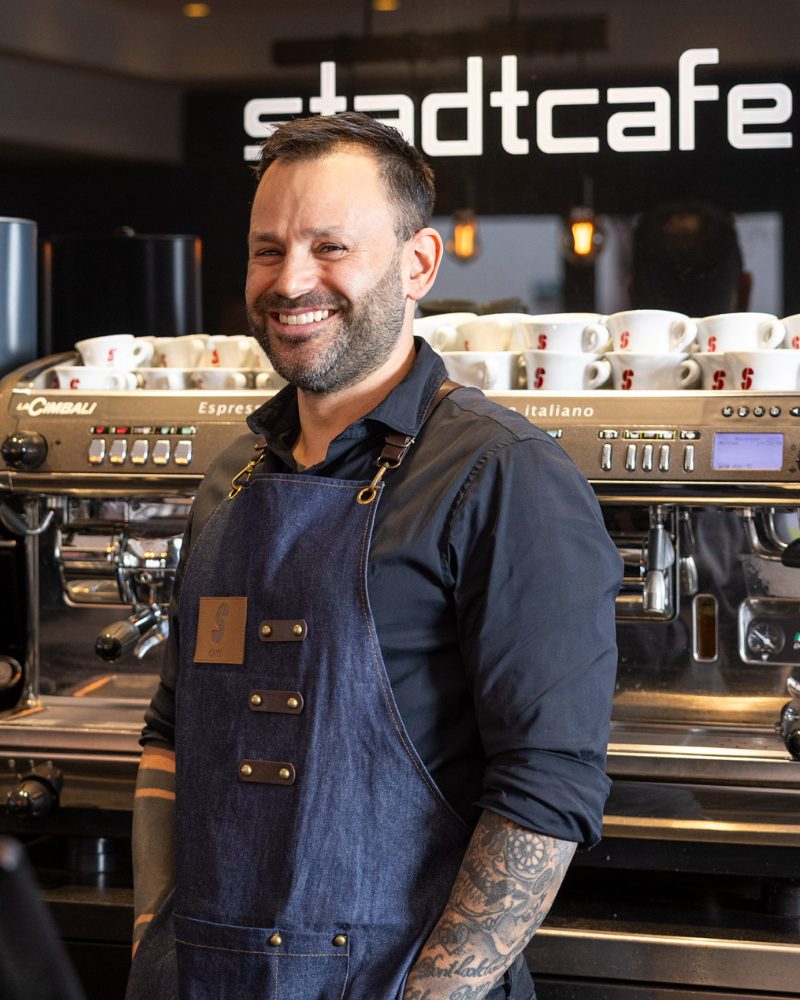 Armin, Barista im Stadtcafé, lächelnd in Jeansschürze vor der Espressomaschine und dem Schriftzug „stadtcafe“