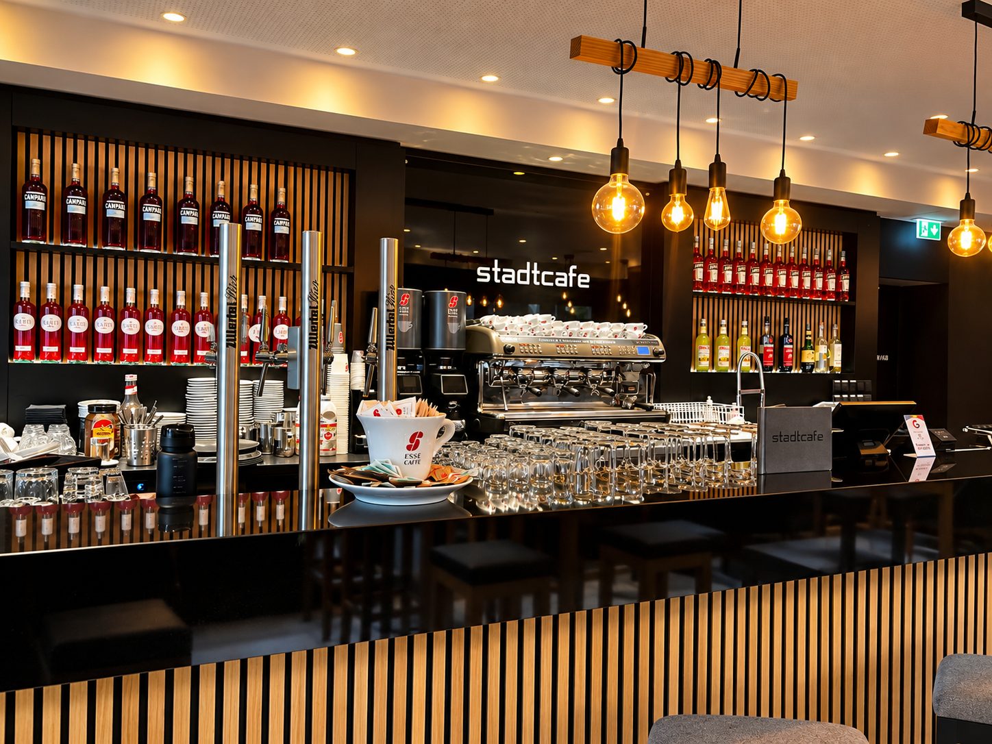 Die Bar mit Zapfhähnen, Gläsern und Espressomaschine unter dem Schriftzug „stadtcafe“, darüber Edison-Birnen