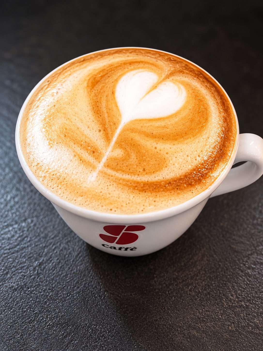Cappuccino mit einem Herz aus Milchschaum in einer weißen Tasse auf dunklem Stein