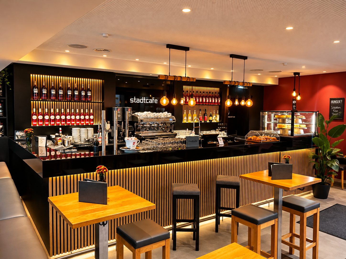 Stehtische mit Hockern vor der Theke, dahinter Espressomaschine, Flaschenwand und die rote Wand