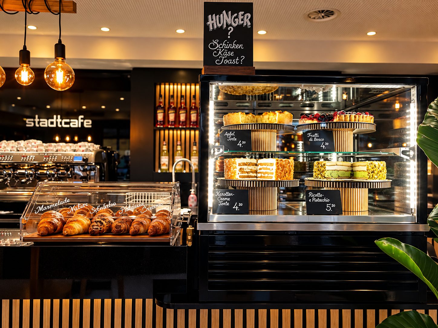 Die Vitrine mit Torten auf Holzetageren, davor Croissants unter der Glashaube, darüber die Tafel „Hunger? Schinken Käse Toast?“