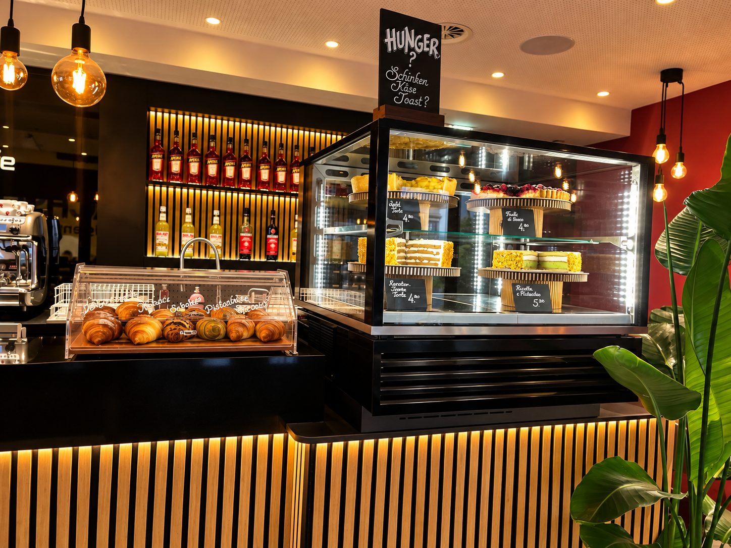 Die Vitrine mit Torten auf Holzetageren, davor Croissants unter der Glashaube und die Tafel „Hunger? Schinken Käse Toast?“